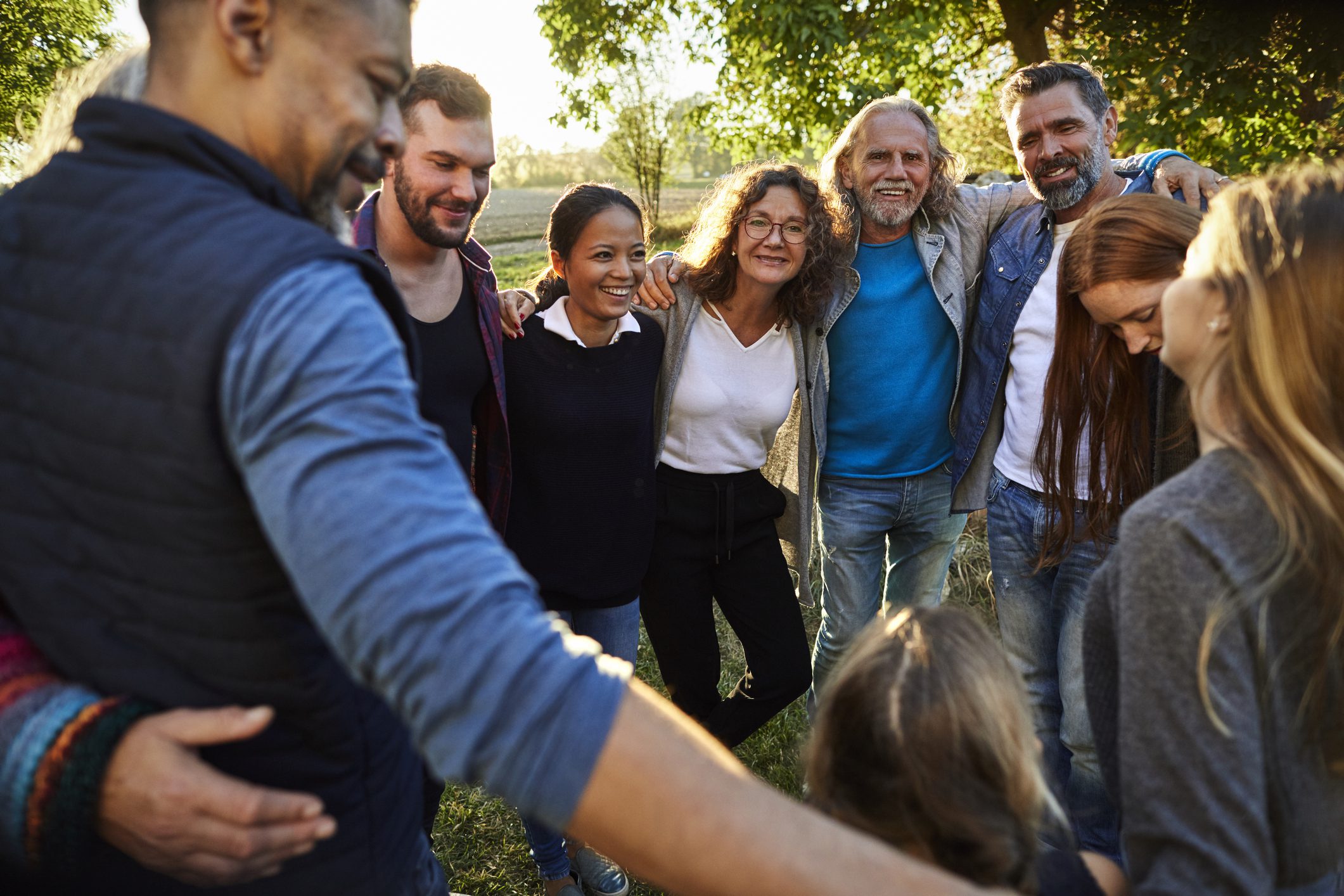 A group of people of all ages stand in a park holding each other in a circle.