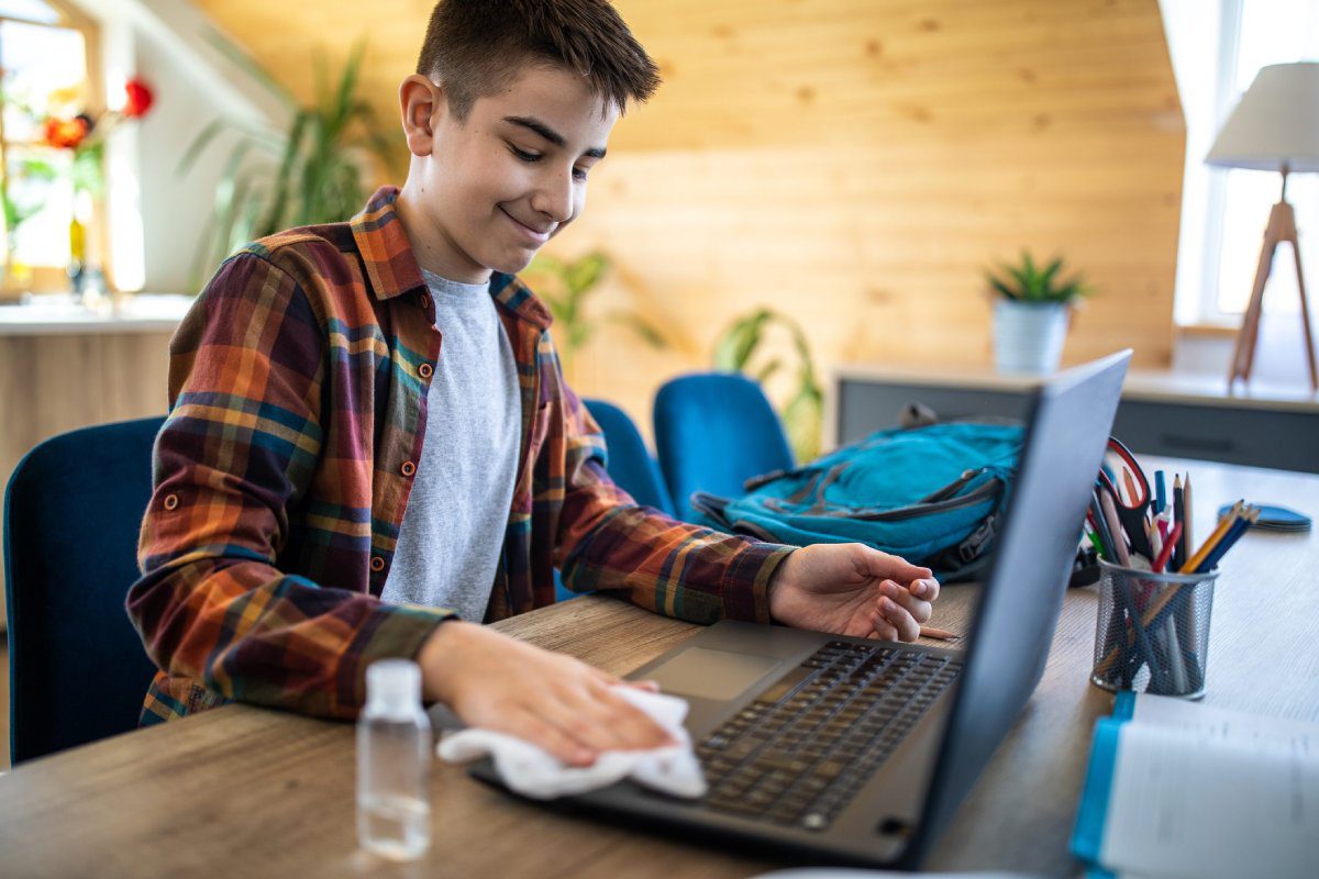 Teen boy cleaning laptop keyboard with cloth at home desk